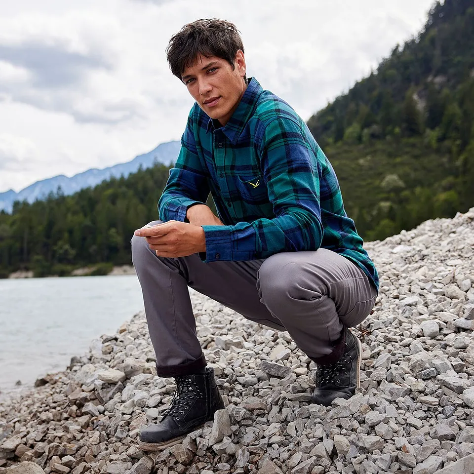 En turgåer i rutete skjote og grå bukse sitter ved elvekanten i et fjellanskap.