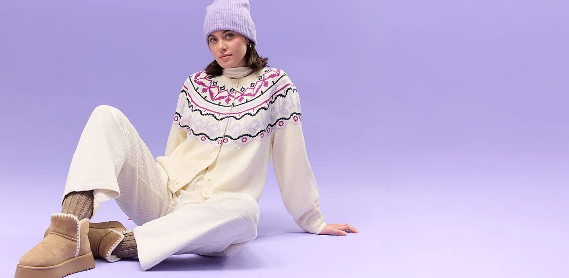 Kremfarget genser med abstrakt koftemønster, hvit bukse og lilla strikkelue.