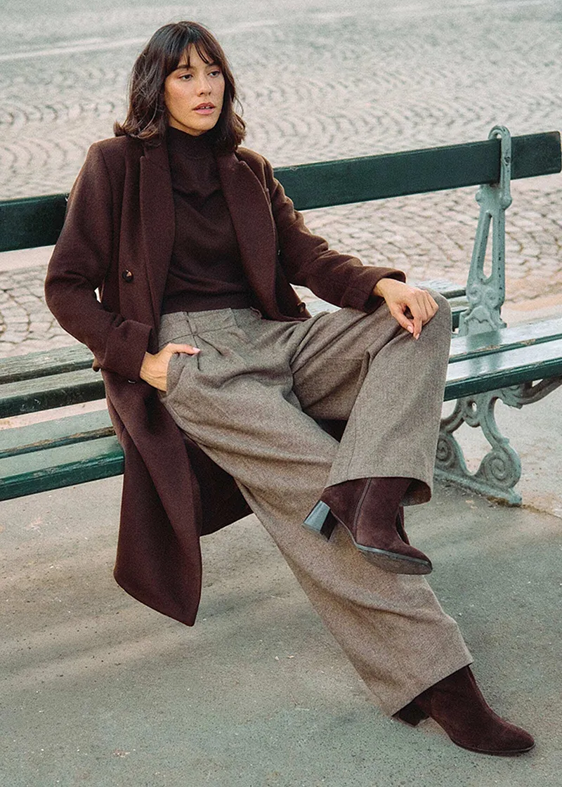 Lang, brun kåpe, høyhalset genser og vid tweedbukse.