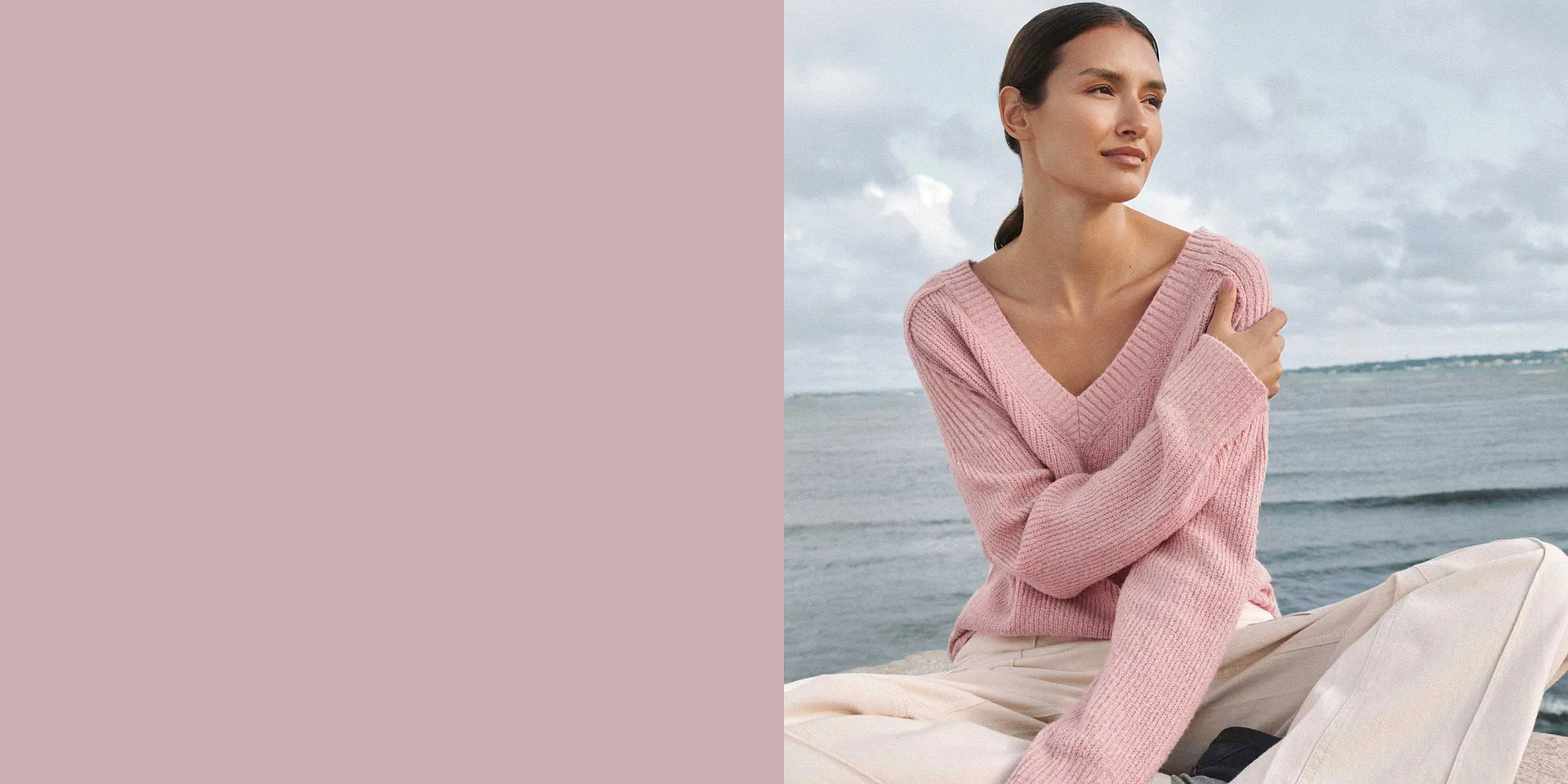 Person i blek rosa, strikket genser med V-hals med havet som bakgrunn.