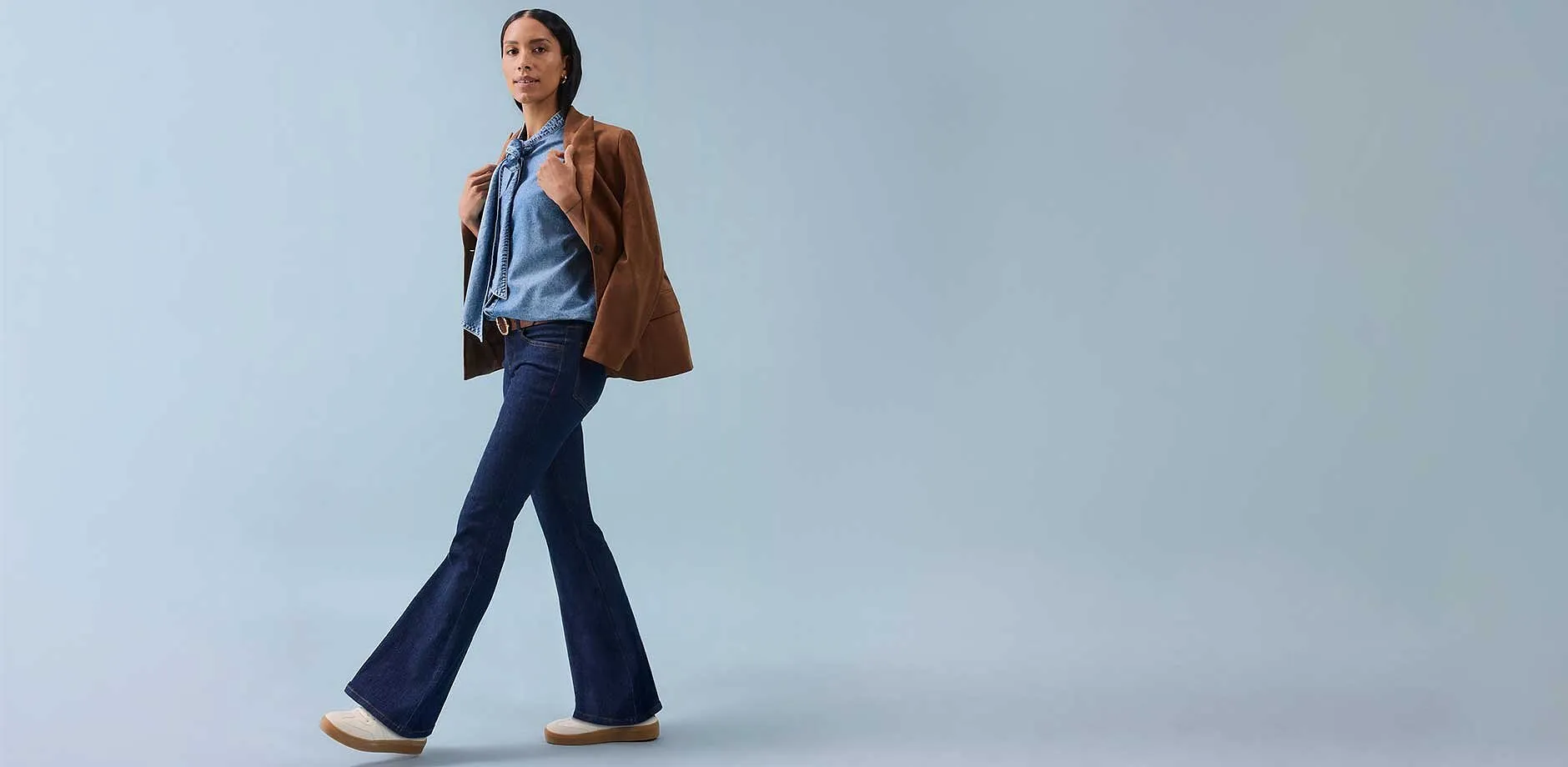 En kvinne i slengbukse, ermel&oslash;s topp og en brun jakke over skuldrene.