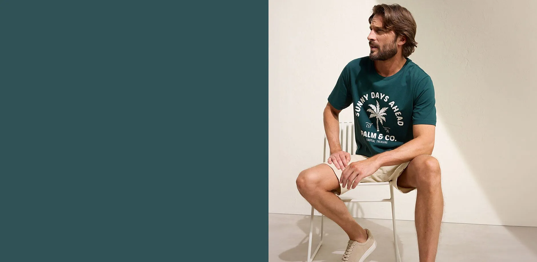 Mann i gr&oslash;nn T-skjorte med palmetrykk, beige shorts og hvite sneakers.