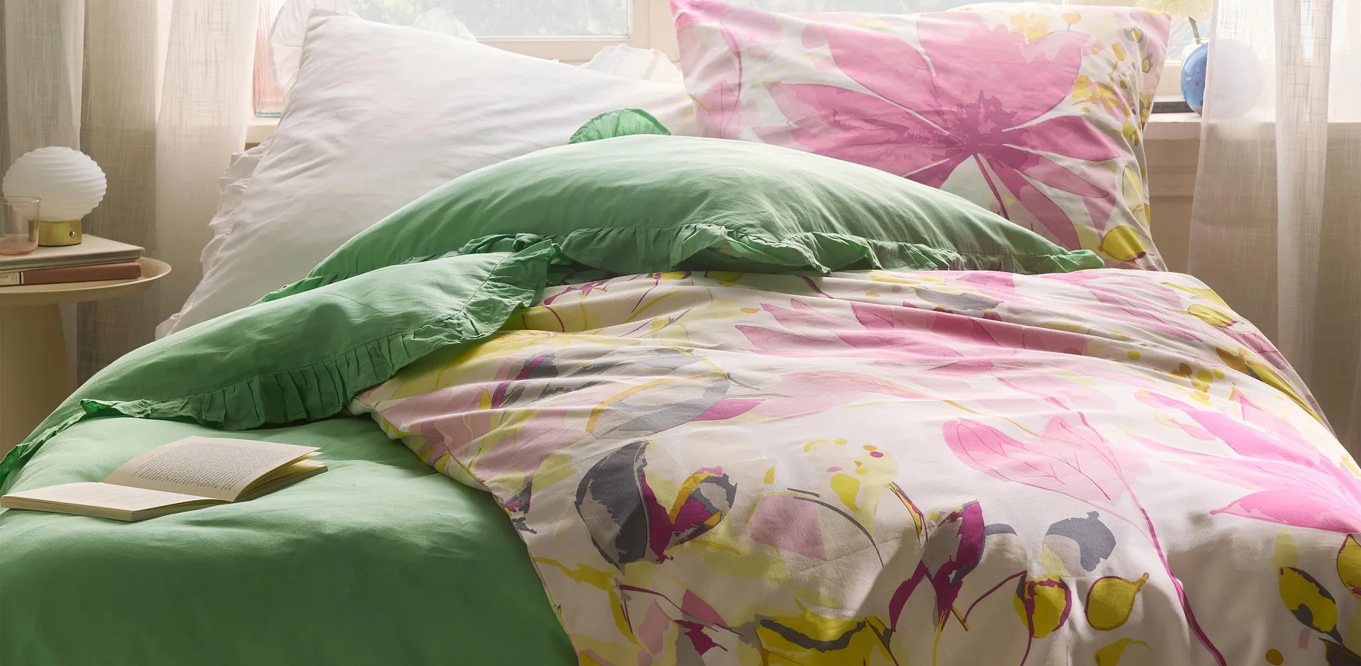 Flerfarget sengesett med blomsterm&oslash;nster kombinert med gr&oslash;nn pute. Lette gardiner rammer inn sengen.