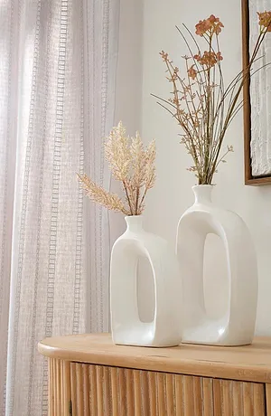 Lyse, luftige gardiner rammer inn elegante hvite vaser med tørkede blomster på et skap i tre.