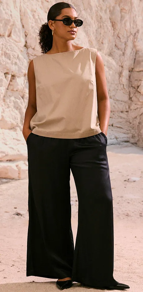 Ermeløs beige topp kombinert med vide, sorte bukser – en elegant look fullført med stilige solbriller.