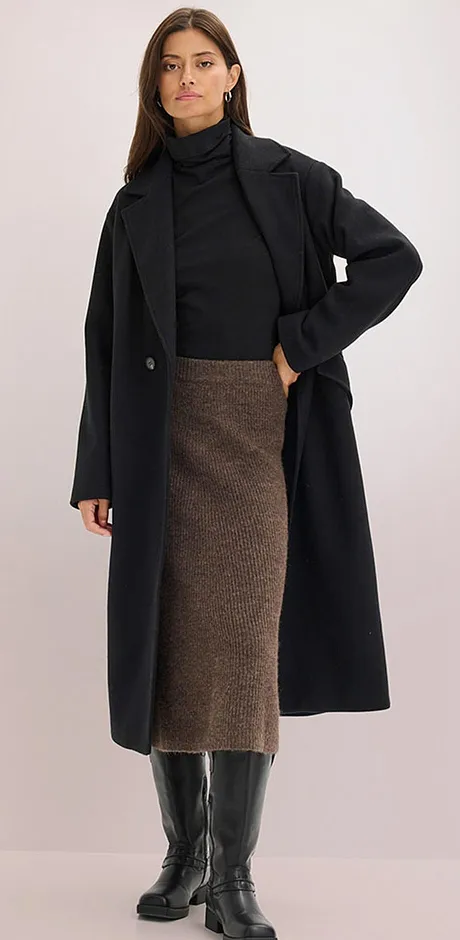 Elegant, sort kåpe over en pologenser kombinert med brunt midiskjørt og knehøye skinnstøvletter.