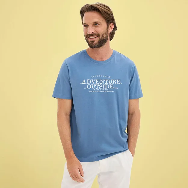 Mann kledd i vid, bl&aring; T-skjorte med hvitt trykk og lys bukse.