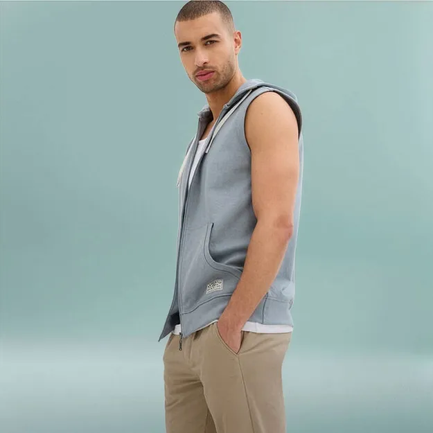 Mann i lysebl&aring; sweat hettevest over hvit shirt og beige bukse.