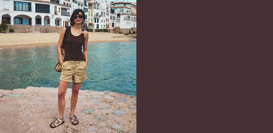 En kvinne bærer en sort singlet og beige shorts med sandaler på stranden.
