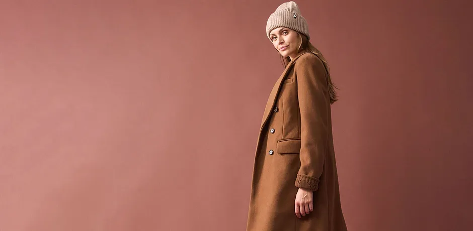 Lang, kamelbrun kåpe, beige, strikket lue og stilrent antrekk under.