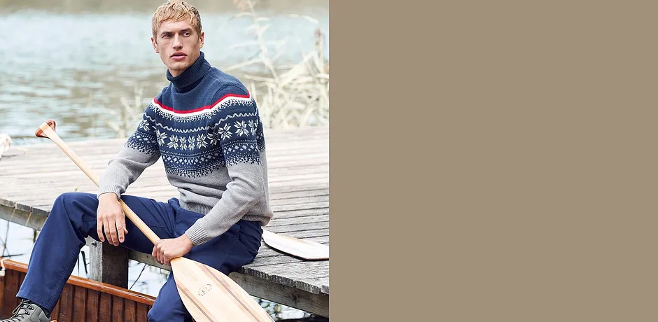 En mann i en genser med koftemønster og jeans sitter avslappet på en båt.