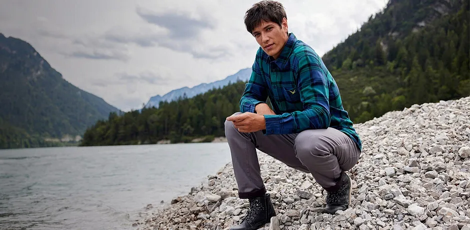 En turgåer i rutete skjote og grå bukse sitter ved elvekanten i et fjellanskap.