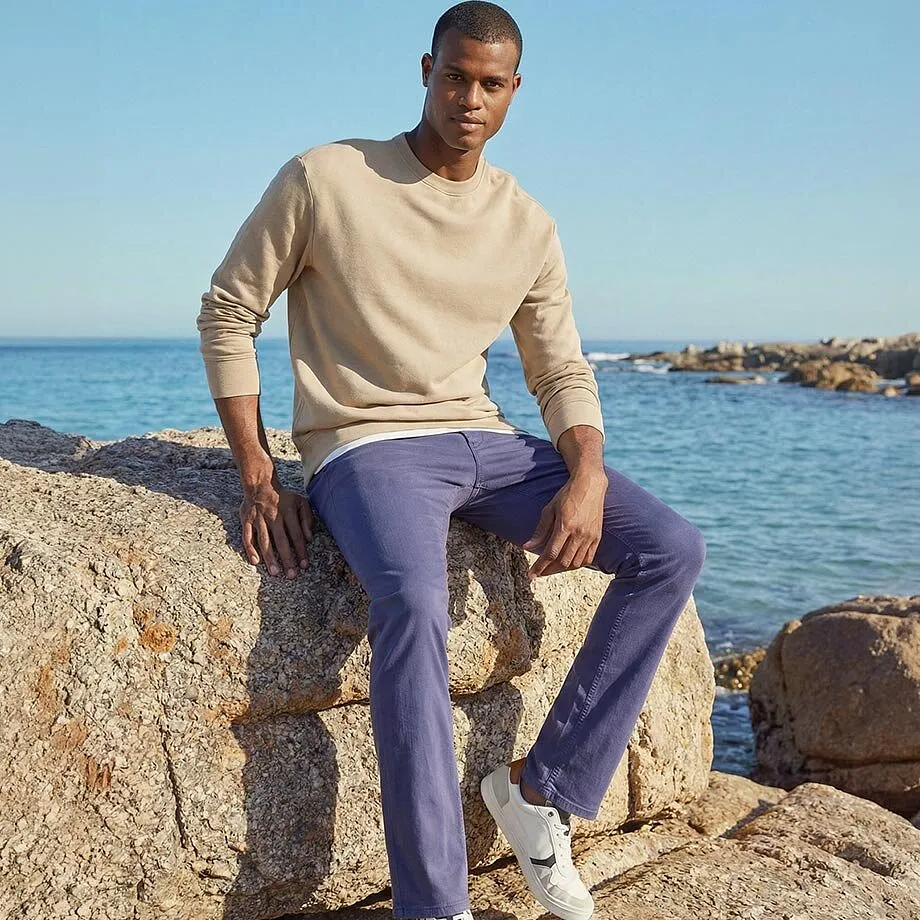 Beige genser og lilla bukse harmonerer perfekt med havet bakenfor.