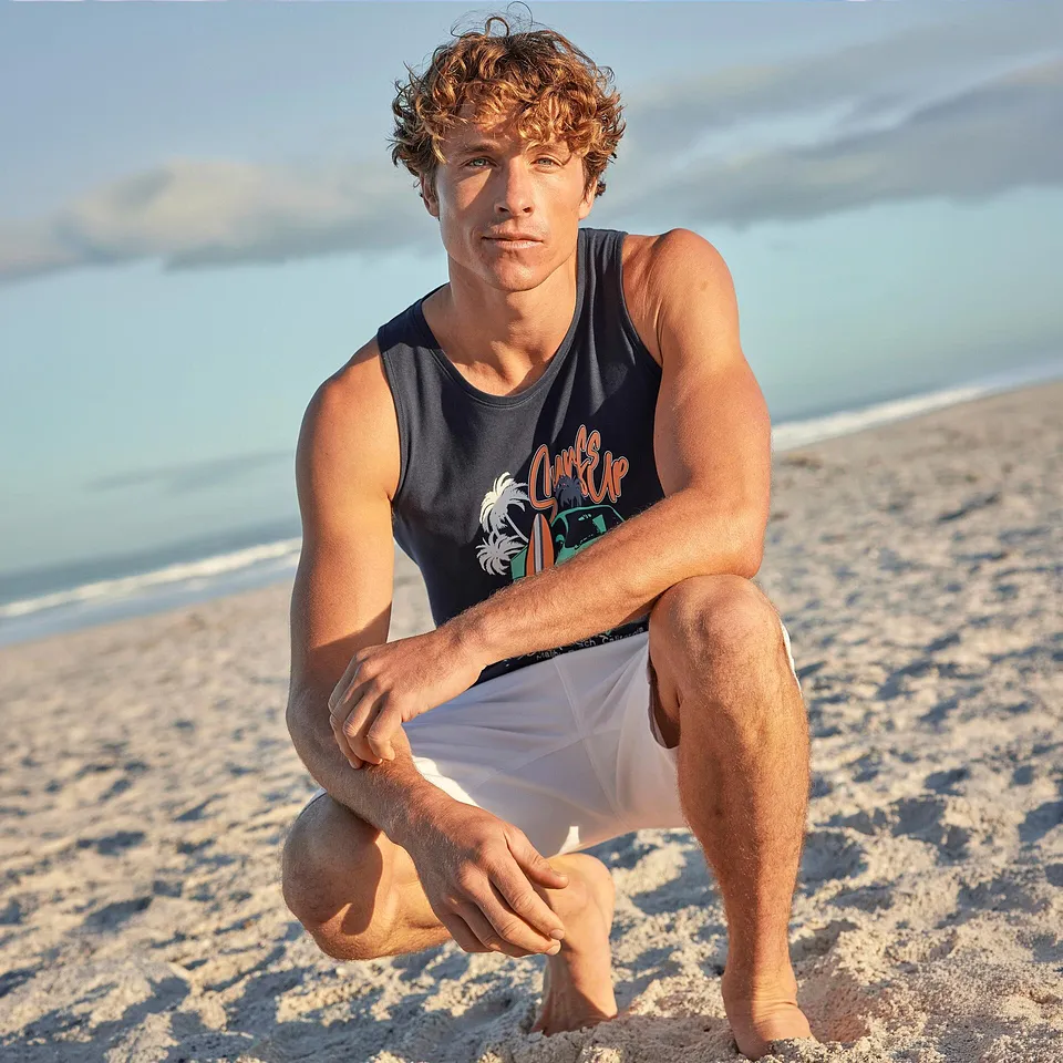 Person i bl&aring; singlet med trykk og hvit shorts p&aring; stranden.