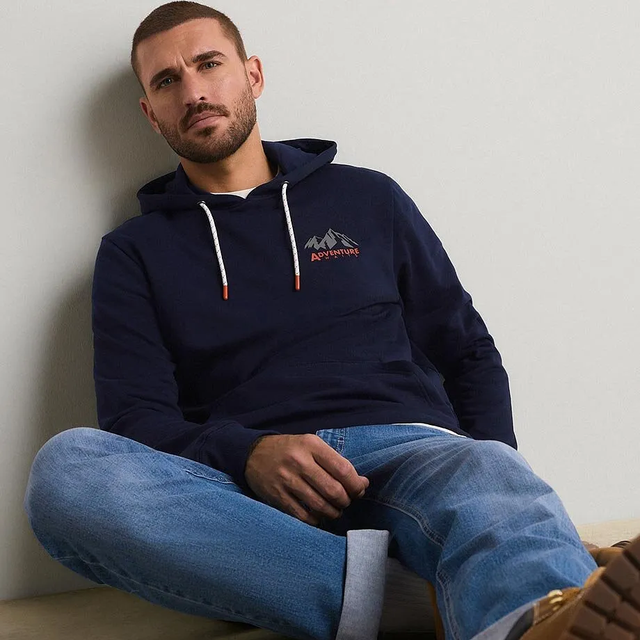 Hen kledd i m&oslash;rkebl&aring; hettegenser med fjellmotiv og avslappede bluejeans.